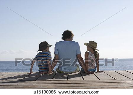 Father and two children (7-9) sitting on jetty back view View Large Photo Image Stock Photograph - Father and two children (7-9) sitting on jetty back view. Fotosearch
