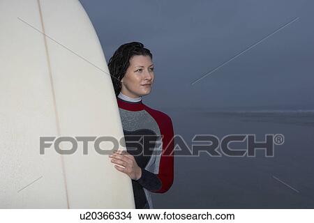 Picture - Female surfer holding surfboard in shallow water at dusk. Fotosearch