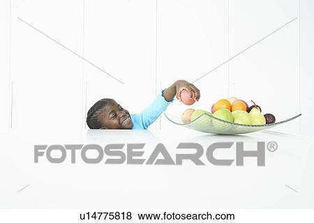 Girl (5-6) standing behind table taking fruit from dish on top View Large Photo Image Stock Photo - Girl (5-6) standing behind table taking fruit from dish on top. Fotosearch