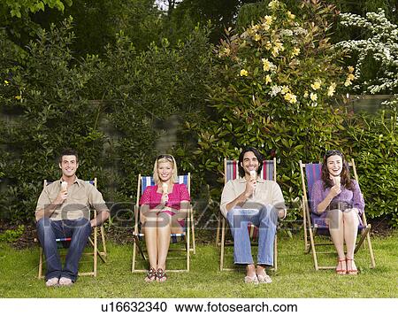 Stock Image - Group of young people sitting in row on deckchairs in garden eating ice creams. Fotosearch