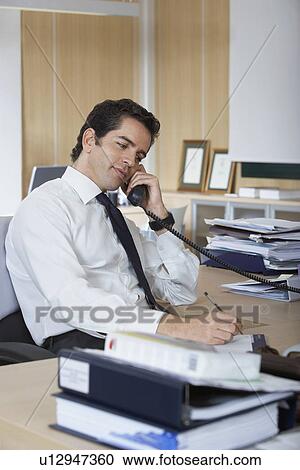 Stock Image - Hispanic businessman using telephone at office desk surrounded by folders. Fotosearch