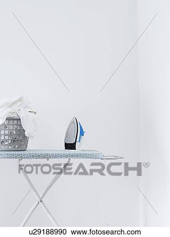 Stock Image - Iron and laundry basket on ironing board against white wall. Fotosearch