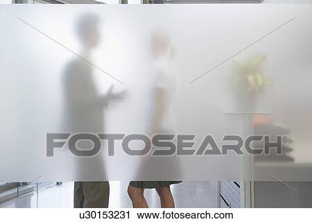 Stock Image - Male office worker talking to female colleague behind translucent wall in office. Fotosearch