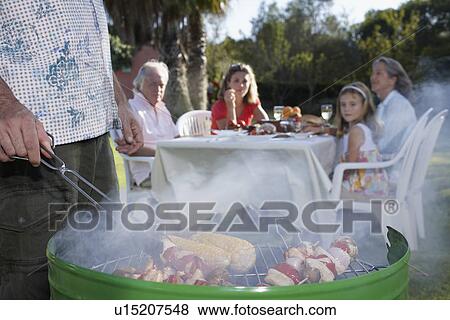 Stock Photo - Man grilling other family members sitting at table in background close-up. Fotosearch