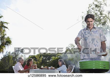 Stock Photography - Man grilling people sitting at table in background. Fotosearch