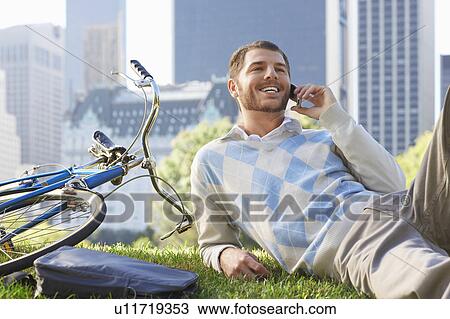 Stock Image - Man lying on lawn in park talking on mobile phone. Fotosearch