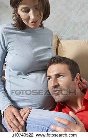 Stock Image - Man putting head on pregnant woman's stomach in living room. Fotosearch