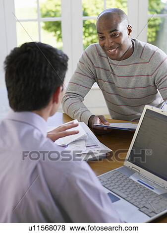 Stock Photo - Man sitting at dinning table with financial advisor using laptop elevated view. Fotosearch