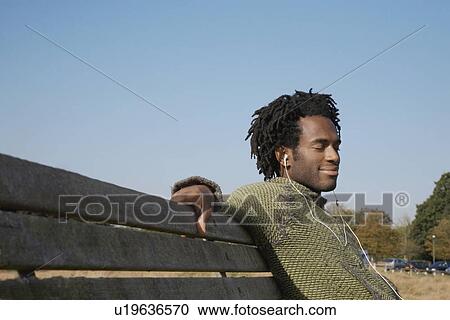 Man sitting on bench listening to music from earphones View Large Photo Image Stock Image - Man sitting on bench listening to music from earphones. Fotosearch