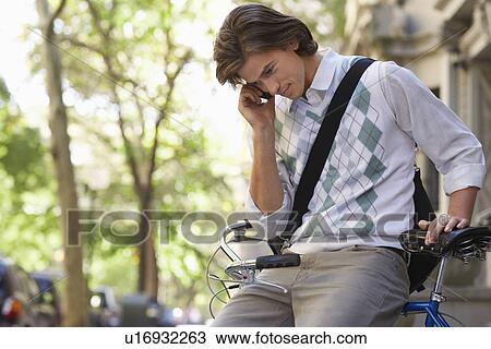 Stock Image - Man sitting on bicycle talking on mobile phone. Fotosearch
