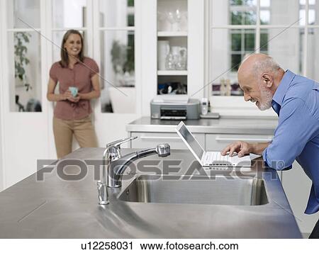 Man using laptop by kitchen sink woman watching from distance View Large Photo Image Stock Image - Man using laptop by kitchen sink woman watching from distance. Fotosearch