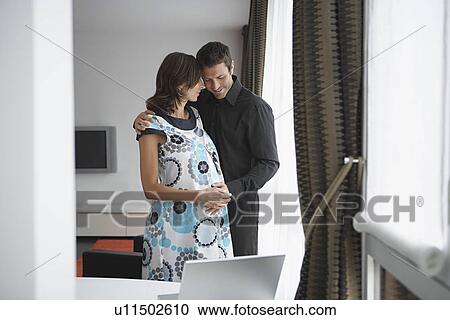 Stock Image - Man with arm around pregnant woman in living room. Fotosearch