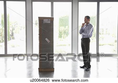 Stock Photograph - Man with hand on chin observing package in empty office. Fotosearch