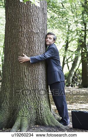 Picture - Mid adult business man hugging tree trunk eyes closed. Fotosearch