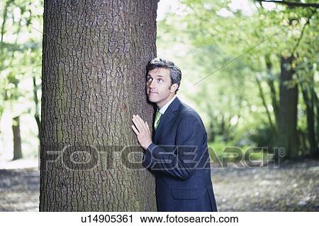 Mid adult business man pressing against tree trunk View Large Photo Image Stock Image - Mid adult business man pressing against tree trunk. Fotosearch