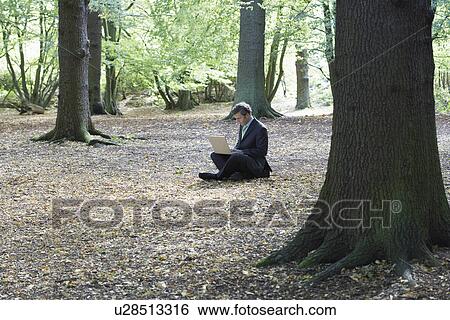 Stock Photograph - Mid adult business man sitting cross-legged in middle of forest working on laptop. Fotosearch