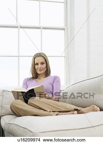 Picture - Mid adult woman reading book on sofa. Fotosearch
