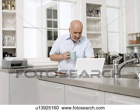 Stock Image - Middle-aged man in kitchen using laptop and holding coffee cup. Fotosearch