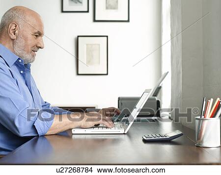 Middle-aged man sitting at desk using laptop profile View Large Photo Image Picture - Middle-aged man sitting at desk using laptop profile. Fotosearch