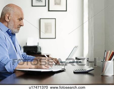 Stock Image - Middle-aged man sitting at desk using laptop and writing in notepad profile. Fotosearch