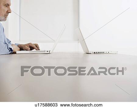 Middle-aged man sitting at desk using laptop another laptop at opposite edge of desk profile View Large Photo Image Stock Image - Middle-aged man sitting at desk using laptop another laptop at opposite edge of desk profile. Fotosearch