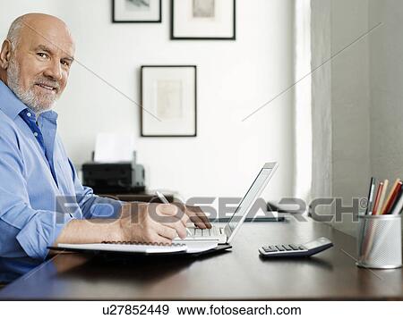 Middle-aged man sitting at desk using laptop and writing in notepad View Large Photo Image Stock Photo - Middle-aged man sitting at desk using laptop and writing in notepad. Fotosearch