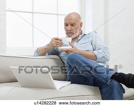 Stock Photo - Middle-aged man sitting on sofa drinking coffee and looking at laptop. Fotosearch