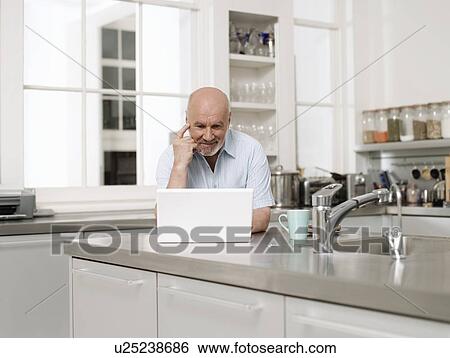 Stock Photograph - Middle-aged man using laptop in kitchen. Fotosearch