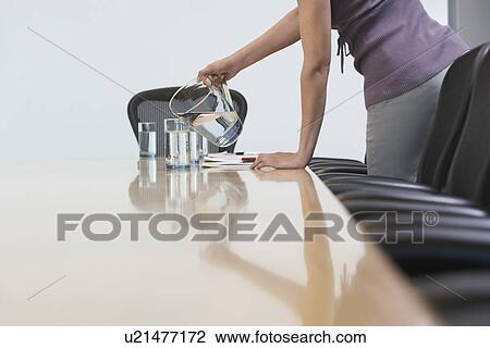 Stock Image - Office worker pouring water into glasses on conference table mid section. Fotosearch