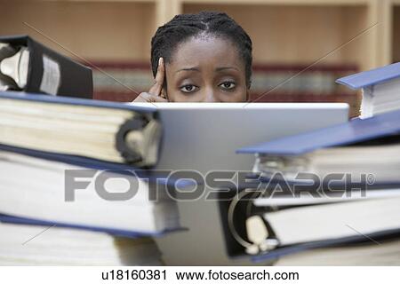 Stock Image - Office worker using laptop sitting behind stacks of documents in office high section. Fotosearch