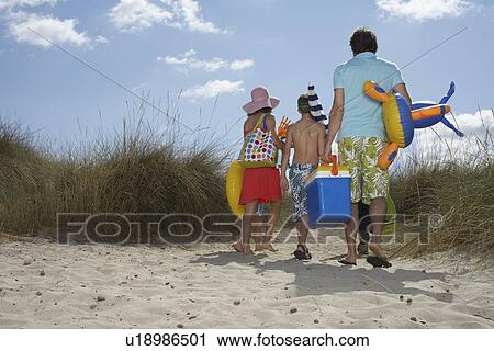 Stock Image - Parents and three children (6-11) carrying beach accessories back view. Fotosearch