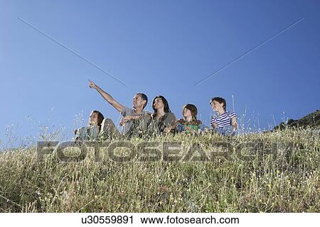 Stock Image - Parents with three children (7-12) sitting on hill man pointing. Fotosearch