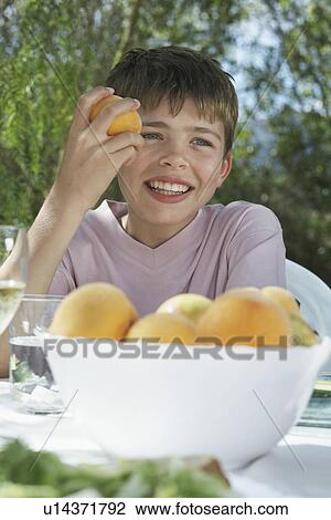 Stock Image - Portrait of boy (10-12) eating peach at garden table smiling. Fotosearch