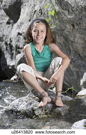 Stock Image - Portrait of girl (10-12) sitting on rock by stream smiling. Fotosearch
