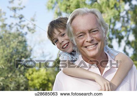 Stock Image - Portrait of grandfather with grandson (7-9) riding piggy back outdoors smiling. Fotosearch