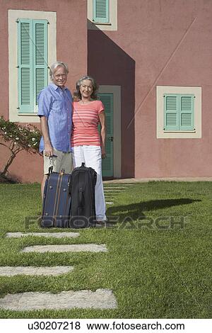 Stock Photo - Portrait of senior couple with luggage in front of house. Fotosearch