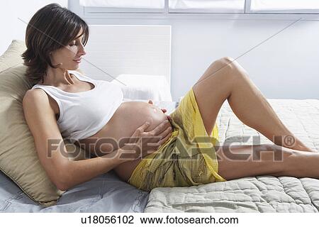 Stock Image - Pregnant woman holding stomach on bed in bedroom. Fotosearch