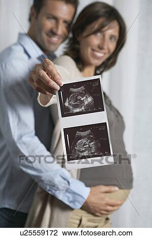 Stock Image - Pregnant woman holding up sonogram with man embracing her from behind indoors. Fotosearch