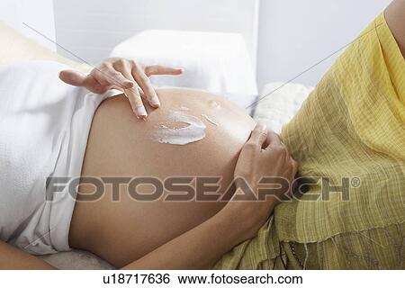 Stock Photograph - Pregnant woman smearing lotion on stomach in bedroom mid section. Fotosearch
