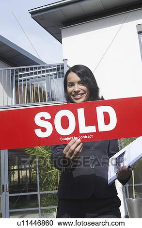 Stock Image - Real estate agent holding sold sign outside house portrait. Fotosearch