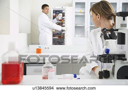 Picture - Scientist using microscope with second scientist getting specimen in laboratory. Fotosearch