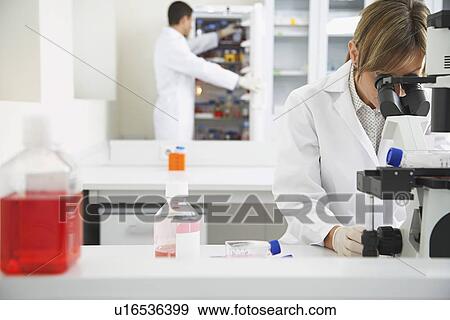 Stock Photo - Scientist using microscope with second scientist looking in refrigerator in laboratory. Fotosearch