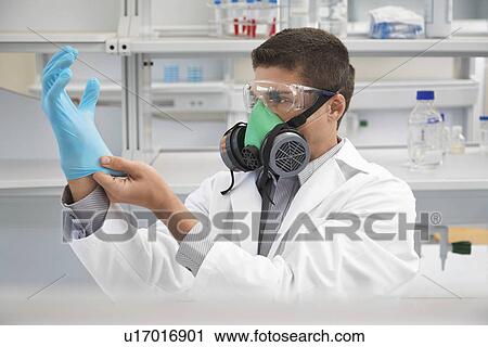 Stock Image - Scientist wearing a gas mask putting on rubber gloves in laboratory. Fotosearch