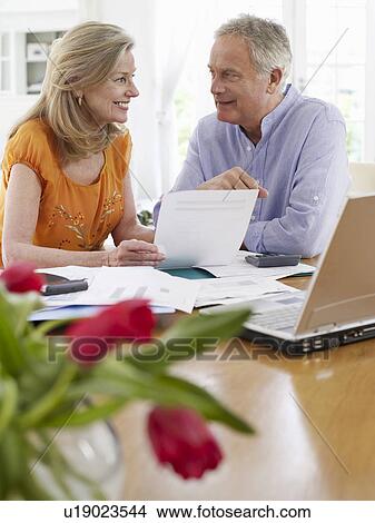 Picture - Senior couple looking at bills sitting at dining table. Fotosearch