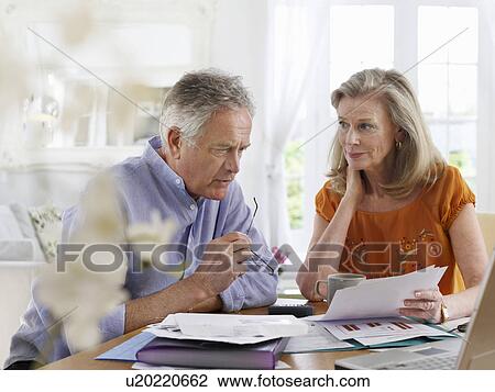 Stock Image - Senior couple looking at bills sitting at dining table. Fotosearch