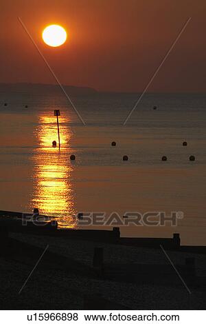 Stock Photo - Sun setting over sea and breakwaters on beach. Fotosearch