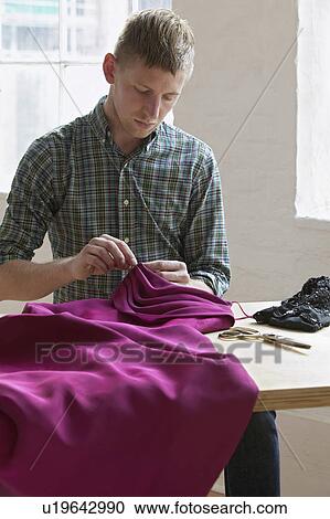 Stock Image - Tailor sewing fabric sitting at table. Fotosearch