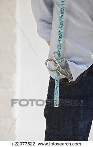 Stock Image - Tailor with scissors in pocket and measuring tape standing indoors mid section close up. Fotosearch