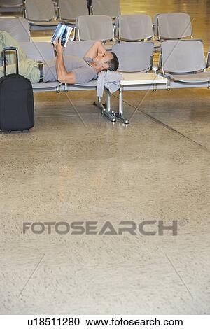 Stock Image - Traveller lying down on chairs reading book in airport lobby. Fotosearch