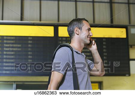 Stock Image - Traveller using mobile phone in front of flight status board in airport. Fotosearch